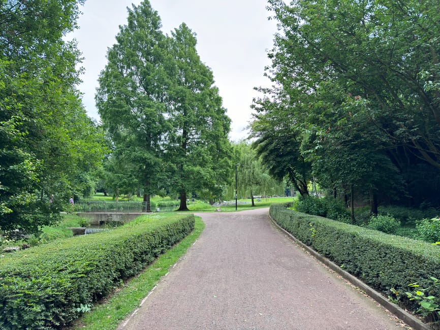 A winding paved pathway through a lush, well-maintained park featuring neatly trimmed hedges along both sides, with a variety of mature trees displaying dense green foliage. The pathway curves gently 