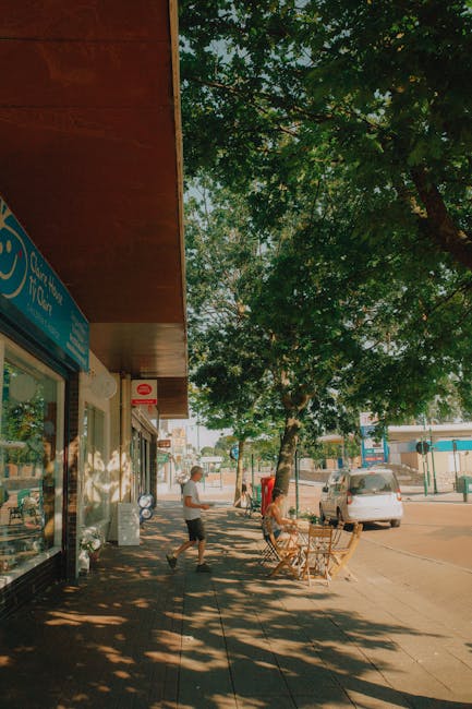 A sunny street scene with a shaded sidewalk lined by a row of lush green trees casting dappled shadows on the pavement. To the left, there is a storefront with a blue sign, part of which is visible, a